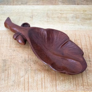 Può includere: Un piatto in legno marrone scuro intagliato a forma di foglia con un gambo e una piccola ghianda attaccata al lato. La superficie del piatto presenta dettagli venati intagliati e una finitura liscia e lucida, creando una ciotola funzionale per piccoli oggetti.