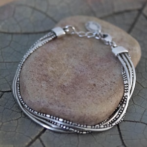 May include: A multi-strand silver-toned bracelet featuring a combination of smooth snake chains and delicate beaded chains. The bracelet includes a metallic clasp mechanism with an adjustable chain extension and a small circular charm at the end.