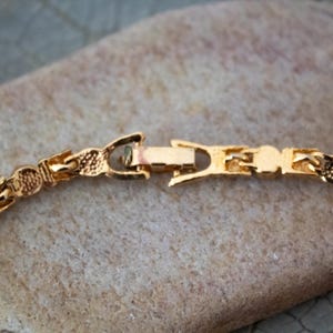 Op de afbeelding: Close-up van een goudkleurige ketting sluiting. De sluiting is rechthoekig met een haak- en lus sluiting. De schakels van de ketting hebben een textuur, patroon ontwerp. De ketting ligt op een lichtbruin, getextureerd oppervlak.