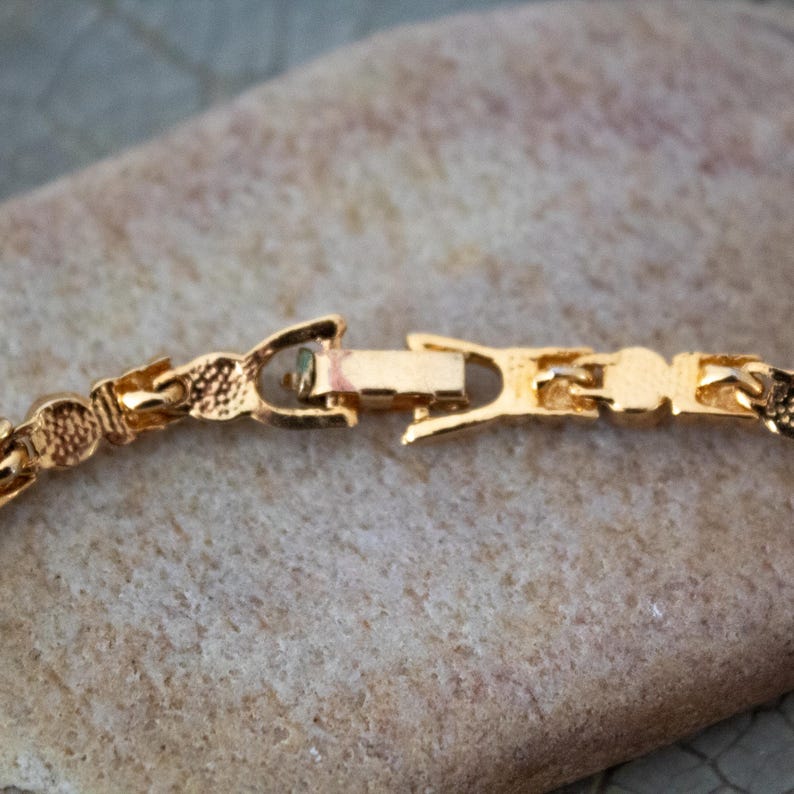 Op de afbeelding: Close-up van een goudkleurige ketting sluiting. De sluiting is rechthoekig met een haak- en lus sluiting. De schakels van de ketting hebben een textuur, patroon ontwerp. De ketting ligt op een lichtbruin, getextureerd oppervlak.