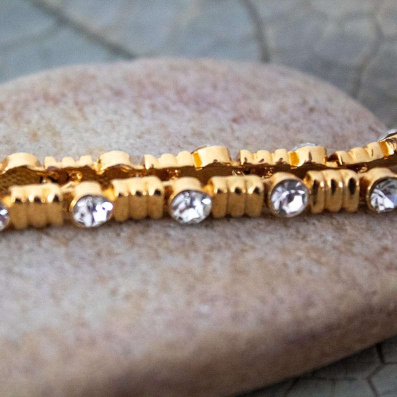 Op de afbeelding: Close-up van een goudkleurige armband met heldere, ronde edelstenen. De armband heeft een textuurontwerp en is geplaatst tegen een neutrale achtergrond, waardoor de glinstering van de edelstenen wordt benadrukt.