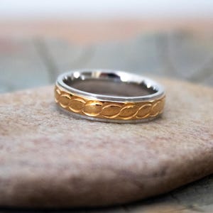Könnte beinhalten: Ein silberner und goldener Ring mit geflochtenem Design. Der Ring hat eine silberne Innenseite und ein goldfarbenes Band mit einem sich wiederholenden Muster aus ineinander verschlungenen Schlaufen. Der Ring liegt auf einem neutralfarbenen Stein.