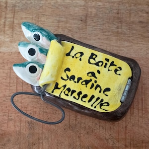 Puede incluir: Una lata de sardinas de cerámica con tres cabezas de pescado. La lata es marrón con una etiqueta amarilla que dice "La Boite à Sardine Marseille" en escritura negra. Se adjunta un lazo de metal.
