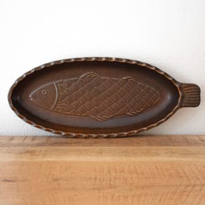 Può includere: Un piatto da portata ovale in ceramica marrone con un design a forma di pesce stampato al centro. Il piatto presenta un bordo smerlato e una superficie strutturata con un motivo a scaglie in rilievo sul corpo del pesce e una coda definita.