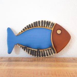 Könnte beinhalten: Ein keramisches Fisch-Wandobjekt mit einem leuchtend blauen Körper, einem einfarbig braunen Kopf mit kreisförmigem Auge und verzierten Flossen mit schwarzen vertikalen Streifen. Das Stück hat eine glatte, glasierte Oberfläche und eine stilisierte Fischform.