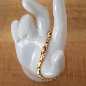 Op de afbeelding: Een goudkleurige armband met heldere, ronde edelstenen. De armband wordt getoond op een witte keramische hand tegen een houten achtergrond. De armband heeft een sluiting en is ontworpen met afwisselende edelsteen- en metalen segmenten.