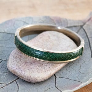 Op de afbeelding: Een zilverkleurige armband met een band van groen, getextureerd materiaal. De armband ligt op een lichtgekleurde steen en een grijs blad, wat het ontwerp en de textuur van de sieraden benadrukt.
