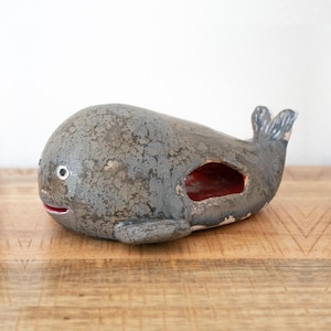 Può includere: Una statuetta di balena in ceramica grigia con una finitura superficiale testurizzata e maculata. La balena presenta un piccolo occhio rotondo con iride bianca e pupilla nera, una sottile bocca rossa e un'apertura laterale che rivela l'interno rosso.