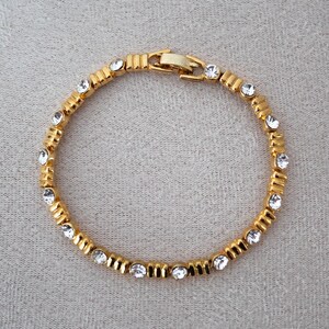 Op de afbeelding: Een goudkleurige armband met heldere, ronde edelstenen. De armband heeft rechthoekige schakels, elk met een edelsteen. De sluiting is eenvoudig. De sieraden worden op een neutrale achtergrond getoond. Lengte: ongeveer 18 cm.