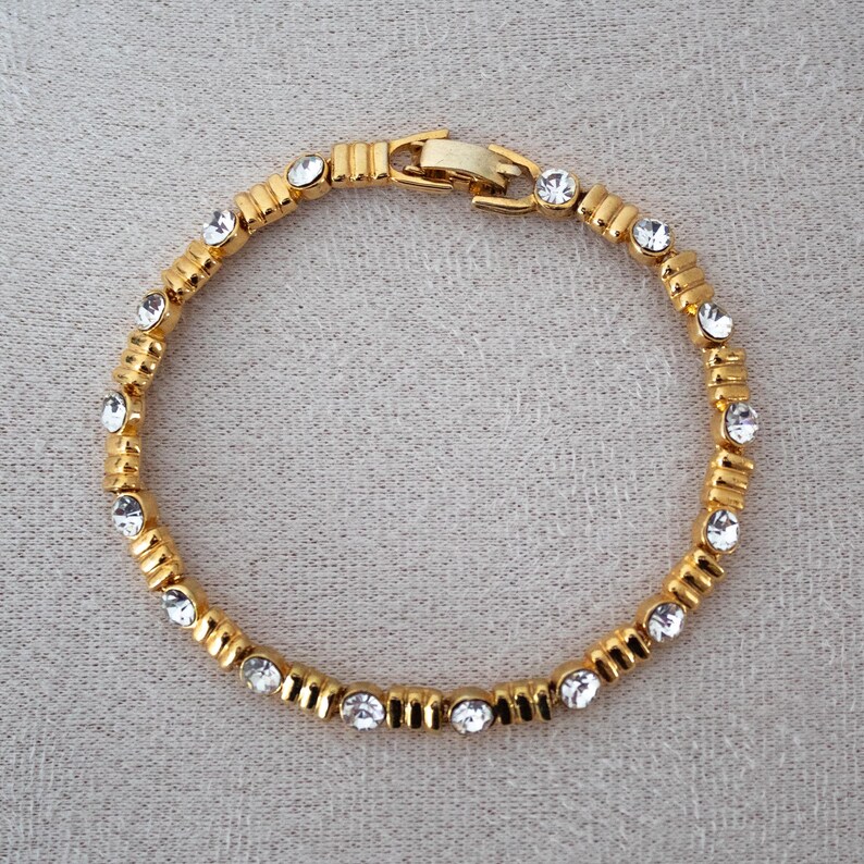 Op de afbeelding: Een goudkleurige armband met heldere, ronde edelstenen. De armband heeft rechthoekige schakels, elk met een edelsteen. De sluiting is eenvoudig. De sieraden worden op een neutrale achtergrond getoond. Lengte: ongeveer 18 cm.