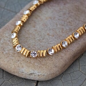 Op de afbeelding: Een goudkleurige ketting met heldere, ronde edelstenen. De ketting ligt op een lichtbruin stenen oppervlak. De edelstenen zijn gelijkmatig verdeeld over de lengte van de ketting, wat een sprankelend effect cre&euml;ert. De ketting is een delicaat sieraad.