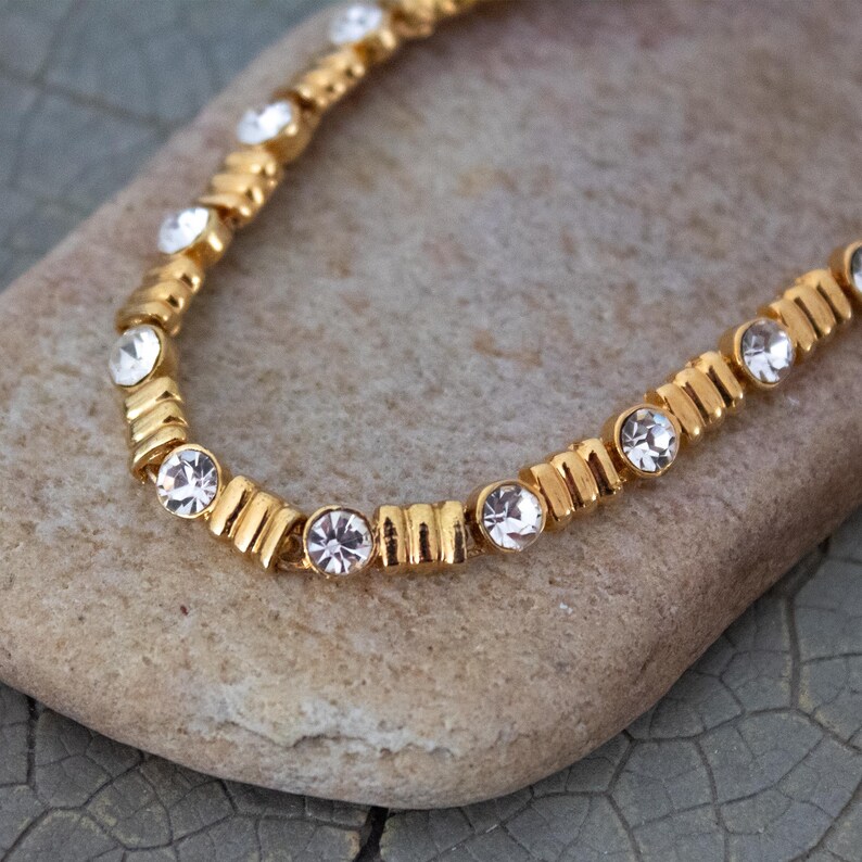 Op de afbeelding: Een goudkleurige ketting met heldere, ronde edelstenen. De ketting ligt op een lichtbruin stenen oppervlak. De edelstenen zijn gelijkmatig verdeeld over de lengte van de ketting, wat een sprankelend effect cre&euml;ert. De ketting is een delicaat sieraad.