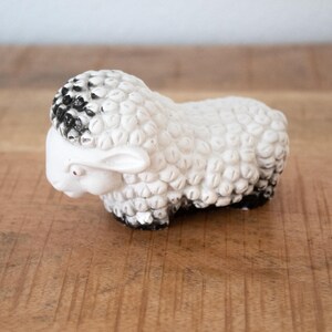 May include: A white ceramic sheep figurine with a textured woolly body and black accents on the head and hooves. The sheep has a small red dot for an eye and is resting on a wooden surface.