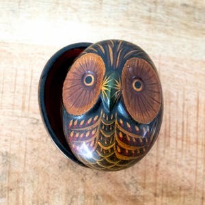 Op de afbeelding: Handbeschilderd houten uilbeeldje met een scharnierend deksel. De uil is voornamelijk zwart met bruine, gouden en rode accenten. De ogen zijn bruin met een lichtere bruine ring rond de pupil. De uil zit in een donkerbruine container.