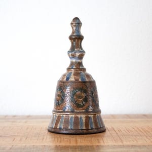 Könnte beinhalten: Handgefertigte Keramikglocke in Braun-, Blau- und Weißtönen. Sie weist ein detailliertes Blumenmuster und vertikale Streifen auf. Die Glocke hat einen verzierten Griff und einen ausgestellten Sockel. Höhe ca. 15 cm.