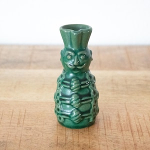 May include: A green ceramic figurine with a face and a hat. The figurine has a mustache and a patterned body. The object is on a wooden surface.