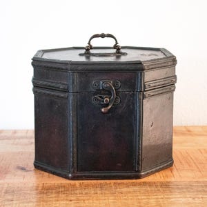 Könnte beinhalten: Eine dunkelbraune, sechseckige Aufbewahrungsbox mit Klappdeckel und einem Metallgriff auf der Oberseite. Die Box verfügt über eine dekorative seilartige Verzierung an den Kanten und einen zentralen Metallverschluss an der Vorderseite.