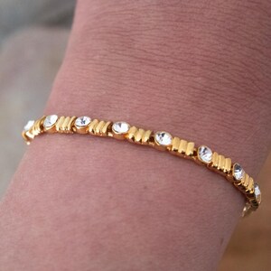 Op de afbeelding: Een goudkleurige armband met heldere, ronde edelstenen. De armband heeft een reeks rechthoekige gouden segmenten, elk gescheiden door een edelsteen. De edelstenen zijn gelijkmatig verdeeld over de lengte van de armband.