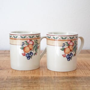 May include: Two cream-colored ceramic mugs with a fruit design. Each mug features a band of tan and green at the top, with a pattern of fruit and leaves. The mugs have a simple, curved handle and sit on a wooden surface.