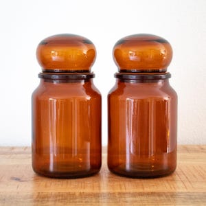 Peut inclure: Deux bocaux d'apothicaire assortis en verre ambré avec des corps cylindriques et des couvercles arrondis en forme de dôme. Le verre est translucide et présente une teinte brune chaude et profonde sur l'ensemble des récipients au fini lisse.