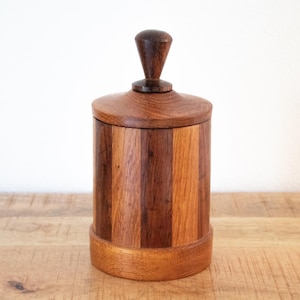 Puede incluir: Un recipiente de madera hecho a mano con tapa y asa pequeña. El recipiente es cilíndrico con rayas verticales de diferentes tonos de madera, incluyendo marrones claros y oscuros. La tapa y la base son de un tono marrón más claro.