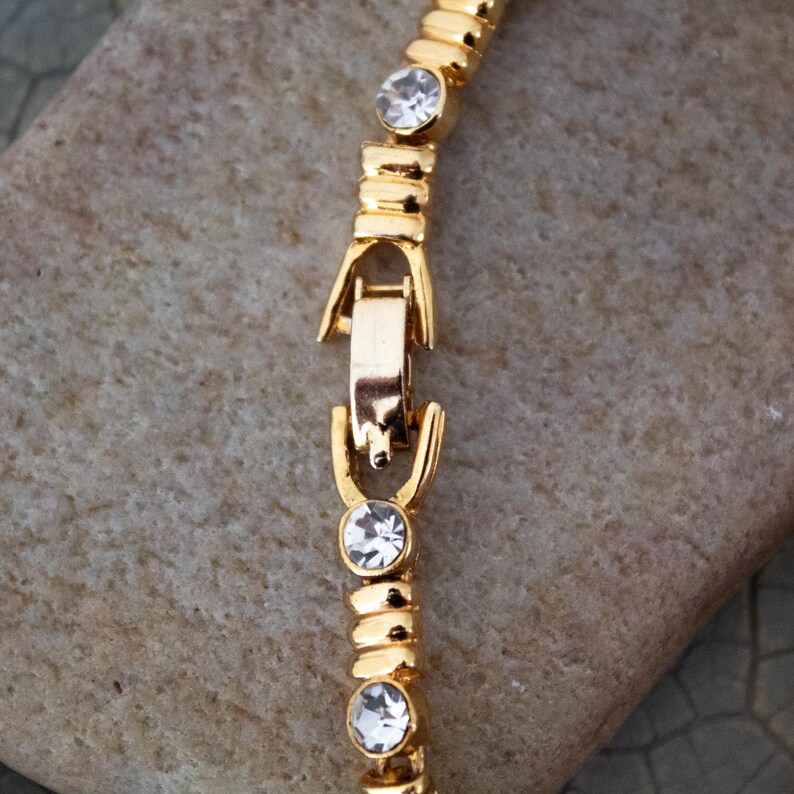 Op de afbeelding: Close-up van een goudkleurige armband met heldere, ronde edelstenen. De armband heeft een sluiting en ligt op een getextureerd, lichtbruin oppervlak. De edelstenen zijn gelijkmatig verdeeld.