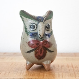 Könnte beinhalten: Eine kleine, gesprenkelte Eulenfigur aus Keramik mit hellgrauer Grundfarbe. Die Eule weist handgemalte Details in Grün, Blau und Dunkelrot auf, darunter ein auffälliges rotes blumenartiges Muster auf der Brust und zwei kleine Füße.