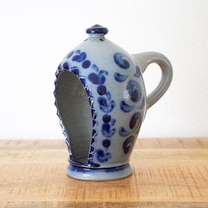 Op de afbeelding: Een keramische kan met een handvat en een grote opening aan de zijkant. De kan is lichtblauw met donkerblauwe bloempatronen en een donkerblauwe rand. De kan staat op een houten oppervlak.
