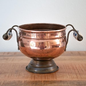 May include: A polished copper bowl with a wide, rounded body and a dark metal pedestal base. The bowl features two curved metal handles on opposite sides, each accented with white ceramic grips decorated with small blue floral patterns.