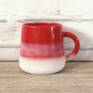 以下が含まれることがあります： グラデーションデザインのセラミックマグ。上部は光沢のある赤色で、まだらなピンクがかった白に変わり、無地の白いベースで終わります。マグカップは丸みを帯びた形状で、お揃いの赤いハンドルが付いています。木製の表面に置かれています。
