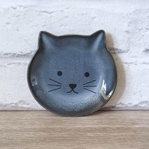 May include: A ceramic cat-shaped dish in a speckled blue-grey glaze. The dish features a cat face with black dot eyes, a nose, and whiskers. The ears of the cat are raised. The dish is on a wooden surface.