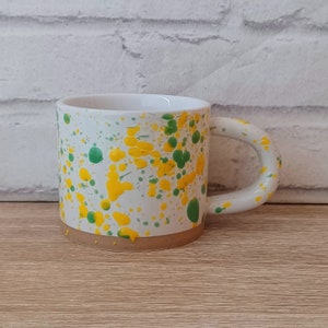 Puede incluir: Taza de cerámica blanca con un diseño moteado en amarillo y verde. La taza tiene un asa redondeada y un borde marrón.