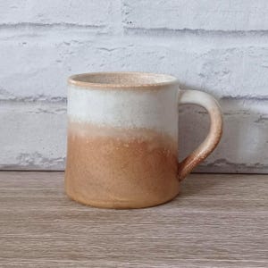 Puede incluir: Una taza de cerámica con la parte superior blanca y un degradado de beige a marrón en la mitad inferior. La taza tiene un asa curva y está colocada contra una pared de ladrillos blancos y una superficie de madera. La taza está diseñada para bebidas calientes.