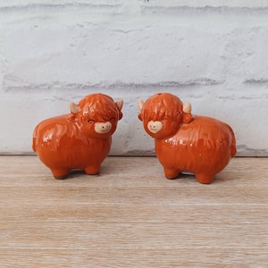 Peut inclure: Deux salières et poivrières en céramique en forme de vaches des Highlands. Les salières et poivrières sont de couleur orange-brun brillant, avec de petites cornes et des visages souriants. Elles sont posées sur une surface en bois clair devant un mur de briques blanches.