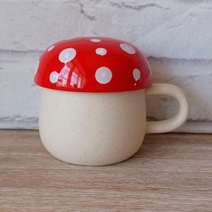 May include: A cream-coloured ceramic mug with a mushroom design. The mug has a red lid with white polka dots. It features a curved handle. The mug is set against a neutral background.