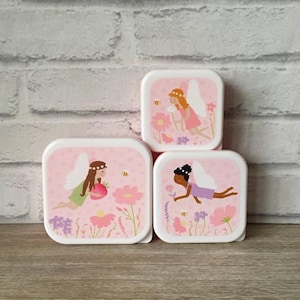 May include: Three pink and white square lunch boxes with fairy illustrations. Each box features a different fairy design with flowers and bees. The boxes are stacked on a wooden surface against a white brick wall.