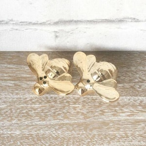 May include: Two gold-colored bee-shaped decorative objects. Each bee has a detailed design with wings and a body. The objects are placed on a light-colored wooden surface, with a white and gray background.