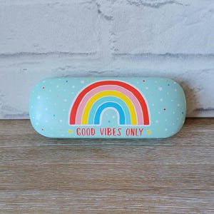 Puede incluir: Un estuche para gafas azul claro con un diseño de arcoíris y las palabras "GOOD VIBES ONLY". El estuche tiene forma rectangular redondeada y está decorado con pequeñas estrellas blancas y confeti colorido.