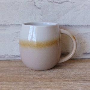 May include: A ceramic mug with a light brown, white and beige ombre glaze. The mug has a rounded body and a handle.