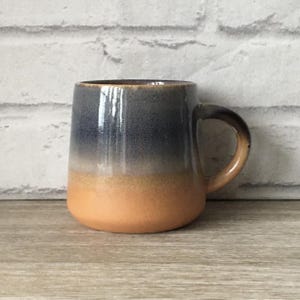 Puede incluir: Una taza de cerámica con un esmalte degradado, que pasa de un azul oscuro a un naranja claro. La taza tiene una forma redondeada y un asa.