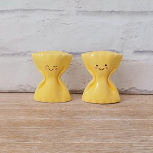 May include: Two yellow ceramic egg cups with smiling faces. The egg cups have an hourglass shape with a ruffled top. The egg cups are on a light brown wooden surface with a white brick background.