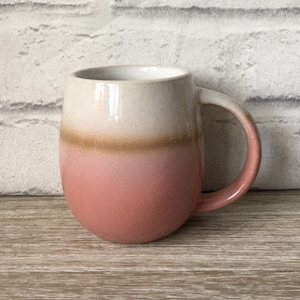 Puede incluir: Una taza de cerámica de color rosa y blanco con un cuerpo redondeado y un asa. La taza tiene una banda marrón en el medio.