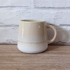 May include: A beige ceramic mug with a rounded base and a handle. The mug features a gradient effect, transitioning from beige at the top to a speckled white at the bottom. The mug is on a wooden surface.