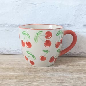 Puede incluir: Una taza de cerámica crema, adornada con un patrón de cerezas rojas y hojas verdes. El borde y el asa son rojos. La taza está diseñada para bebidas calientes.
