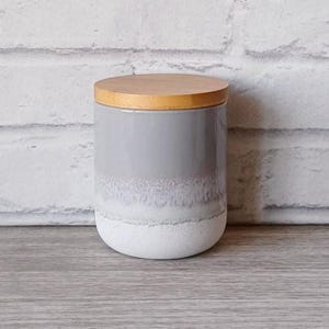 Puede incluir: Un tarro cilíndrico de cerámica con un esmalte gris claro que se desvanece a blanco en la base. Tiene una tapa de madera natural. El tarro está sobre una superficie de madera, con una pared de ladrillo blanco al fondo.