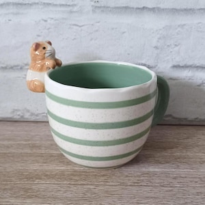 May include: A ceramic mug with horizontal green stripes and a green interior. A small, detailed hamster figurine is attached to the rim. The mug has a rounded shape and a green handle. The background is a white brick wall.