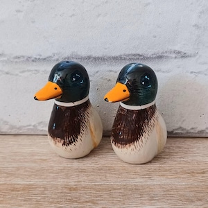 Puede incluir: Un par de saleros y pimenteros de cerámica con forma de pato. Los patos tienen cabezas de color verde oscuro, picos naranjas y cuellos blancos. Los cuerpos son marrones y blancos. Los saleros y pimenteros se muestran sobre una superficie de madera clara.