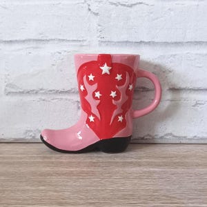 May include: A pink ceramic mug in the shape of a cowboy boot. The boot features a red overlay with white star details. The handle and body are pink, with a black heel. The mug is designed to resemble a Western-style boot, a fun novelty item.