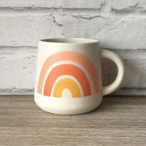 May include: Cream-colored ceramic mug with a rounded base and a large handle. The mug features a rainbow design in shades of orange and yellow. The mug is set against a wooden surface and a white brick wall.