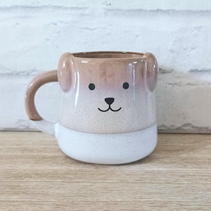 Puede incluir: Taza de cerámica con forma de cara de perro, con asa. La taza presenta una base blanca, una sección media moteada marrón claro y blanca, y orejas marrones claras. La cara del perro tiene ojos de puntos negros y una boca negra curvada.